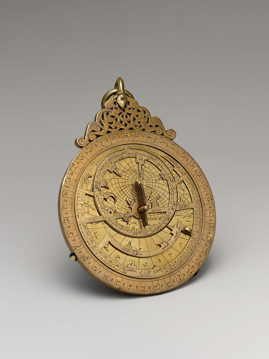 Front of a circular brass-colored astrolabe with an engraved outer scale, a pierced inner framework, a central pin and a suspension ring above. This photograph does not show the maker's inscription on the reverse.