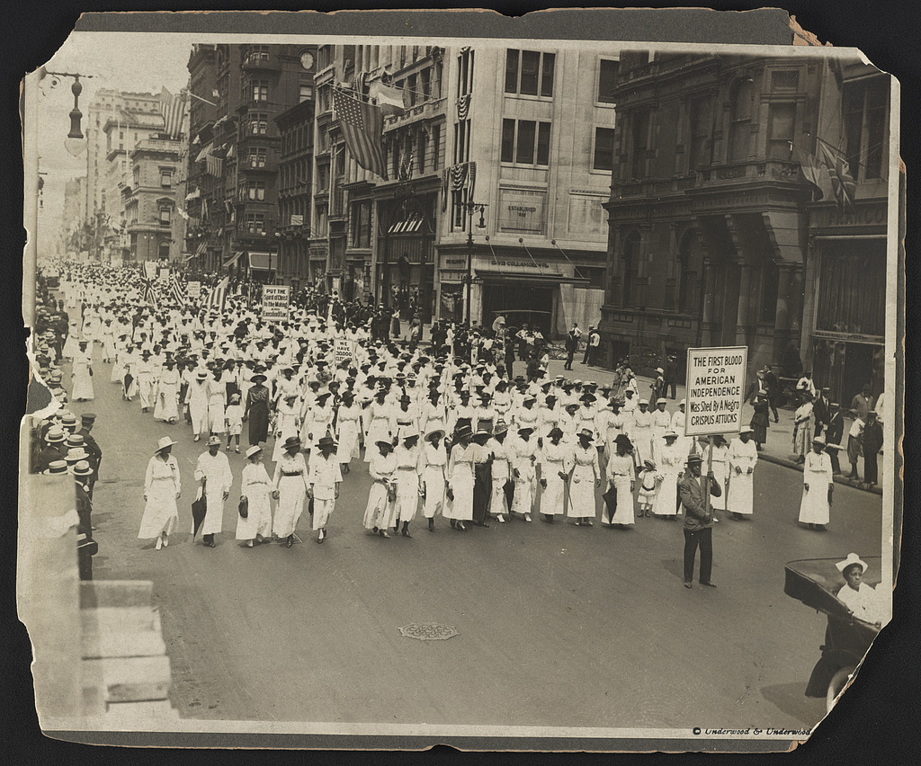 An elevated view along a city street, bordered by an irregular surviving mount. A procession in rows, many in light clothing and hats, fills the street; placards, spectators, flags and tall buildings are visible. A right-foreground placard invokes American independence and Crispus Attucks. Sound, complete attendance and policy outcomes cannot be observed here.